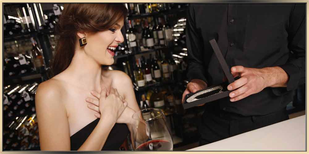 A beautiful young woman with brown hair up, wearing a black evening dress, is being surprised when she sees her jewelry gift. She's holding her hands together at her chest and smiling at the gorgeous necklace in the black jewelry box that is being presented to her. There is a glass of wine on the table in front of her as well. This picture is a great reflection of the ARY D'PO's article about jewelry as a gift.