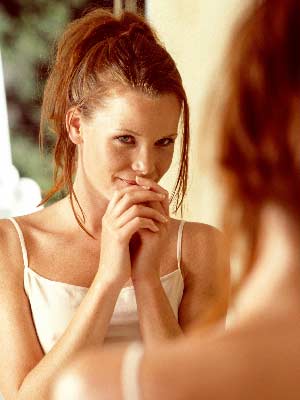 A young woman with red hair and white summer top is looking at a mirror.