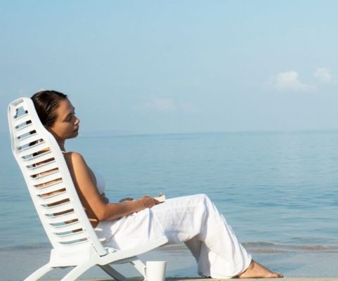 a woman wearing white beach clothing relaxing on a white chair on the beach 