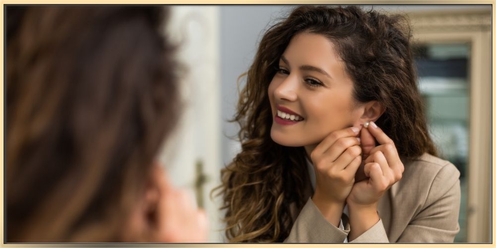 A beautiful woman with curly long dark hair is putting on earrings while looking into mirror and smiling