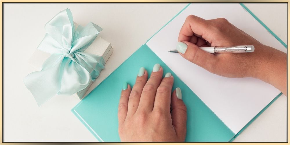 arydpo presents a white jewelry box with a Tiffany blue ribbon on a white surface and a greeting card of the same colors. There are two hands, one hand is on one side of the card and another one has a pen implying that the person is ready to write a gift message. The nails on the hands match the theme colors of white and Tiffany blue.