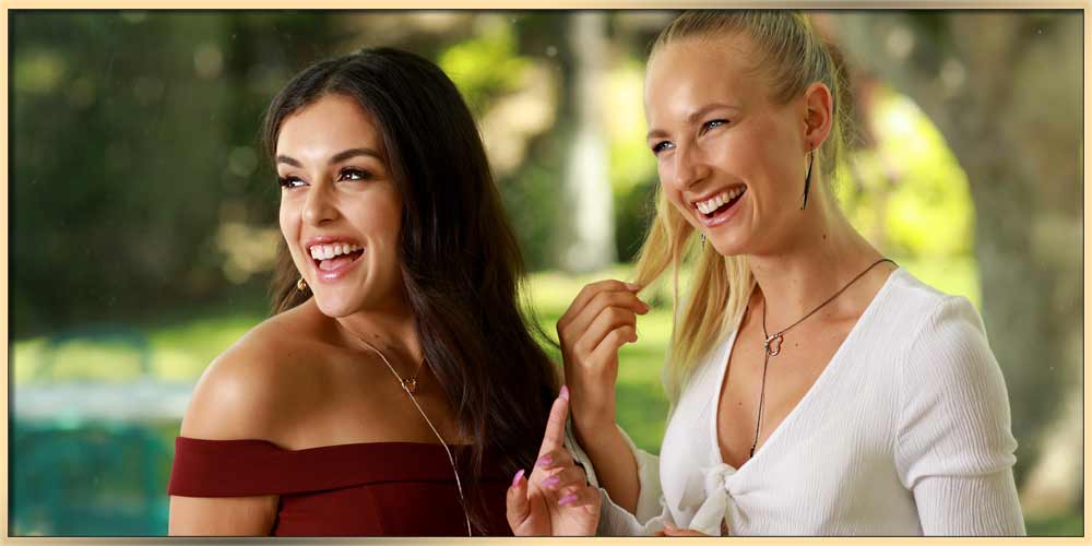 Two young women are looking on a side while smiling. One has dark hair and wearing dark ruby color dress, and the other one has light hair and is wearing white dress - she is touching her hair in a cute manner. Both are wearing arydpo heart necklaces.