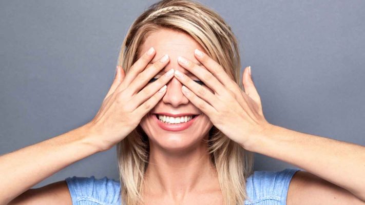 A young woman with blond hair wearing a light blue top is smiling while closing her eyes with her hands