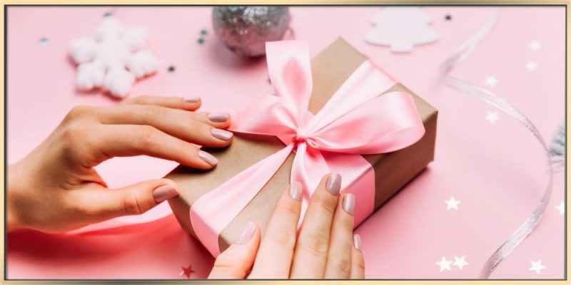 A woman's hands with clean beautifully done nails finishing wrapping a holiday gift while surrounded by a holiday ornaments.