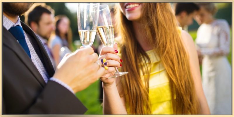 Wedding guests are socializing. A woman with long blond hair is wearing a yellow dress. A man in a suite and the woman hold champagne glasses. They look happy.